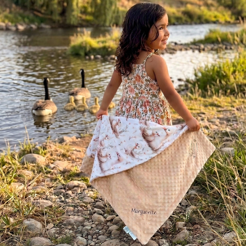 A young girl stands by a riverbank at sunset holding up a quilted blanket with a floral print side and a beige patterned reverse side. Two geese are swimming in the water behind her, with trees and grass in the background.
